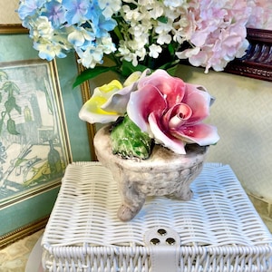 May include: A porcelain floral arrangement featuring three roses in shades of pink, yellow, and white. The roses are arranged in a small, rustic-looking pot with three legs. The arrangement is sitting on a white wicker basket.