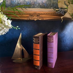 May include: A brass sailboat sculpture and two vintage Reader's Digest Condensed Books are displayed on a dark wooden surface. The books have colorful spines and text. The background features a framed painting and floral arrangements.