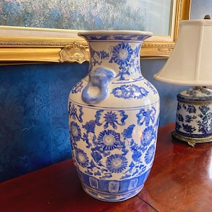 May include: A large, white and blue ceramic vase with a floral pattern. The vase has a flared rim, a cylindrical body, and a blue elephant-shaped handle. The vase is decorated with blue floral motifs and is displayed on a wooden surface.