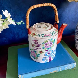 May include: A white ceramic teapot with a red spout and handle, adorned with floral patterns in pink, purple, and green. The handle is made of woven rattan. The teapot sits on a blue book.