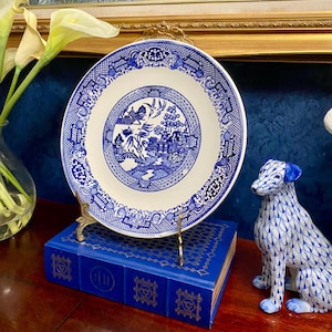 May include: A blue and white porcelain plate with a detailed landscape design, displayed on a gold-colored stand atop a blue book. A blue and white dog figurine is to the right. The plate has a detailed border.