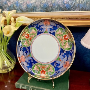 May include: A decorative ceramic plate with a gold rim, featuring a blue background with floral and leaf patterns in green, red, and pink. The plate is displayed on a gold stand, resting on a green book titled "Basic Botany".