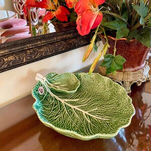 May include: A green ceramic serving dish shaped like a cabbage leaf with a smaller bowl attached to the side. The dish is on a wooden table.