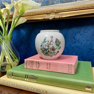 May include: A small, white ceramic vase with a floral design in pink, green, and blue sits atop a stack of vintage books. The books are in shades of pink, green, and cream. A glass vase with white flowers is in the background.