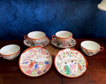 Könnte beinhalten: Set aus vier japanischen Vintage-Teetassen und Untertassen mit Blumen- und Geisha-Motiven. Die Tassen und Untertassen sind weiß mit goldenen Verzierungen und zeigen farbenfrohe Blumenmuster und Bilder von Geishas.