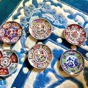 May include: Six decorative ceramic plates with intricate floral and geometric designs. The plates feature a variety of colors, including red, blue, green, and gold. The plates are arranged on a blue and white patterned rug.