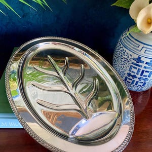 May include: An oval silver-toned metal serving platter featuring a polished finish and a decorative rope-style border. The center of the platter is embossed with a raised, stylized branch or antler design that extends across the surface.