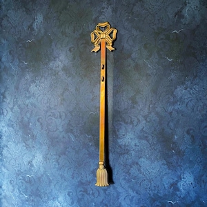 May include: A decorative, gold-colored wall hanging with a bow at the top and a tassel at the bottom. The long, rectangular piece has three small circular cutouts. The background is a dark blue wall with a subtle floral pattern.