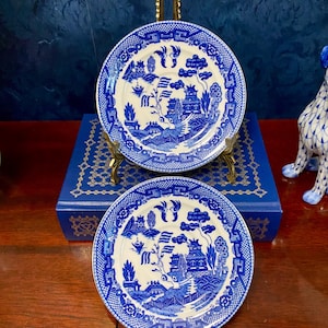 May include: Two blue and white ceramic plates with a Willow pattern design. The plates are displayed on gold-colored stands, showcasing a classic, detailed landscape scene. The plates are approximately 8 inches in diameter.