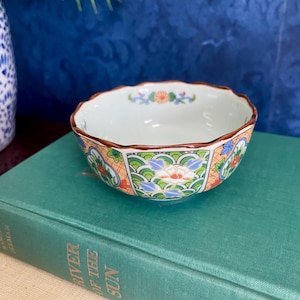 May include: A small, white ceramic bowl with a scalloped rim and a colorful floral design. The bowl is decorated with orange, green, blue, and white flowers and leaves. The bowl is sitting on top of a green book.