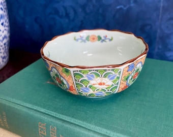 May include: A small, white ceramic bowl with a scalloped rim and a colorful floral design. The bowl is decorated with orange, green, blue, and white flowers and leaves. The bowl is sitting on top of a green book.
