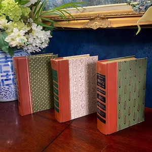 May include: Four vintage books with decorative covers. The spines are a reddish-orange color, and the covers feature patterns in green and white. The books are arranged on a dark wooden surface, with a vase of flowers in the background.