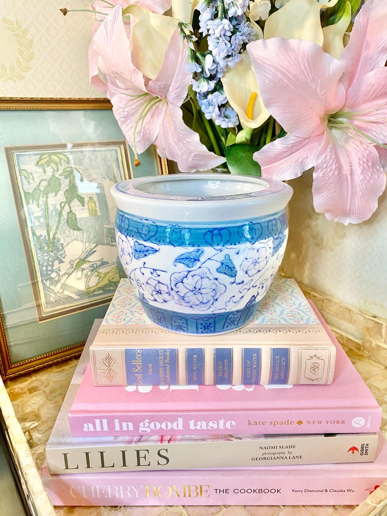 May include: A stack of four books with pink and white covers. The top book is titled "all in good taste" with the author "kate spade" and the location "NEW YORK". The second book is titled "LILIES" with the author "NAOMI SLADE" and the photographer "GEORGIANNA LANE". The third book is titled "CHERRY BOMBE THE COOKBOOK" with the authors "Kerry Diamond & Claudia We". A blue and white ceramic planter sits on top of the books.