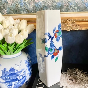 May include: White ceramic vase with a geometric shape and a floral design featuring blue, red, and green leaves and fruit. The vase is sitting on a black surface with a white fluffy object in the background.