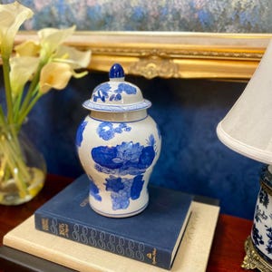 May include: A white and blue ceramic ginger jar with a lid, featuring floral and grape cluster designs. The jar is placed on a stack of books, with a lamp and flowers in the background. The jar is a decorative home accent.
