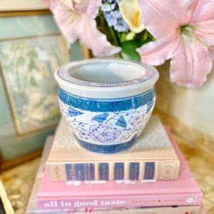 May include: A blue and white porcelain planter with a floral design sits on top of a stack of books. The books are titled "all in good taste", "LILIES", and "CHERRY BOMBE THE COOKBOOK".