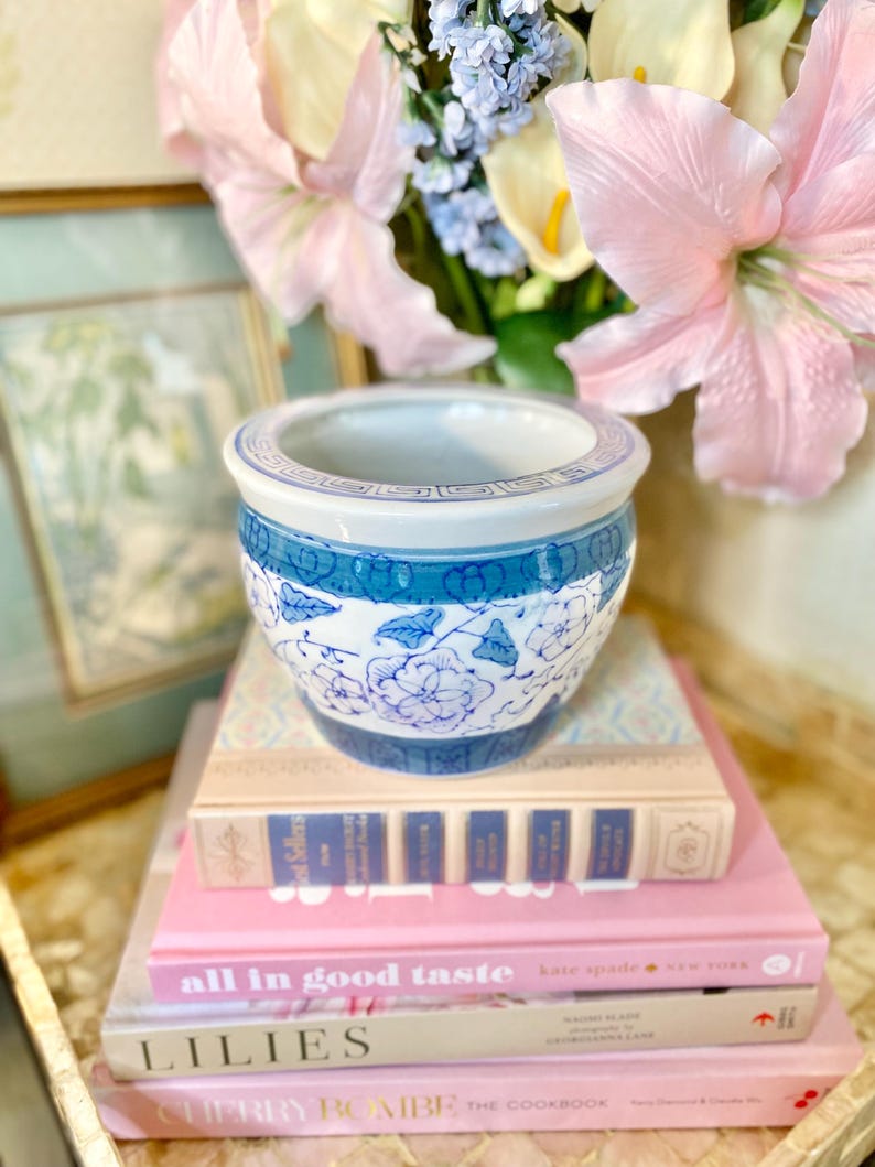 May include: A blue and white porcelain planter with a floral design sits on top of a stack of books. The books are titled "all in good taste", "LILIES", and "CHERRY BOMBE THE COOKBOOK".