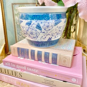 May include: A blue and white ceramic planter with a floral design sits on top of a stack of four books. The books are all pink and have titles that include "all in good taste", "LILIES", "CHERRY BOMBE", and "THE COOKBOOK".