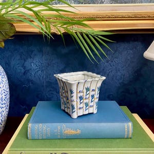 May include: A square, white ceramic planter with a blue and gold floral design sits atop a stack of books. The planter has a flared rim and is supported by small feet. The books are various colors, including blue and green, and have gold lettering.