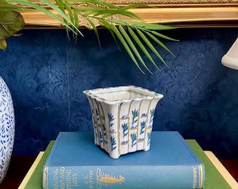 May include: A square, white ceramic planter with a blue and gold floral design sits atop a stack of books. The planter has a flared rim and is supported by small feet. The books are various colors, including blue and green, and have gold lettering.