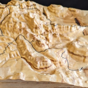 May include: A detailed, three-dimensional wooden topographical map, showcasing a landscape with varying elevations. The map is crafted from light-colored wood, with darker wood grain patterns. Black lines delineate specific geographical features.