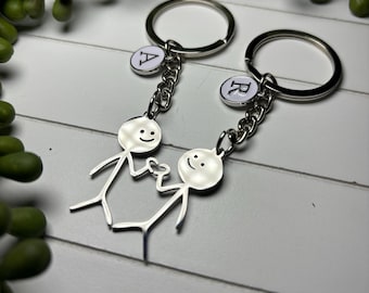 May include: Two silver keychains with stick figure charms holding hands. Each keychain has a round ring, a chain, and a small charm with a letter. The background is a white wooden surface with green plant accents.