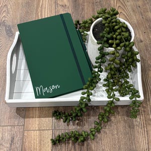 Może przedstawiać: Zielony, ciemny notes z napisem "Mason" w kolorze białym na okładce. Notes leży na białej tacy z zieloną sukulentem.