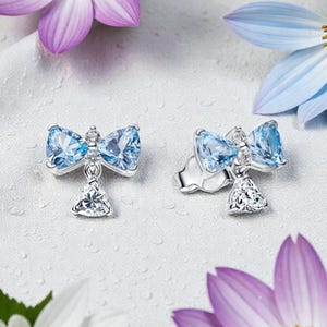 Może przedstawiać: Srebrne kolczyki w kształcie kokardki z jasnoniebieskimi kamieniami szlachetnymi. Każdy kolczyk ma górę w kształcie kokardy i wiszący trójkątny klejnot. Kolczyki są na białym tle z fioletowymi, białymi i niebieskimi kwiatami.