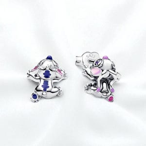 Pu&ograve; includere: Un paio di orecchini a lobo argentati con un design a personaggio dei cartoni animati. Gli orecchini hanno dettagli in smalto blu, rosa e viola. Sono esposti su uno sfondo bianco.