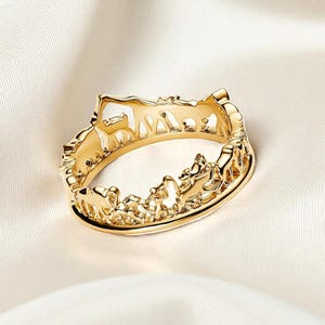 May include: A gold ring with a detailed, openwork design. The ring features a continuous pattern of stylized figures, possibly animals or people, creating a unique and artistic look. The ring is set against a soft, white background.