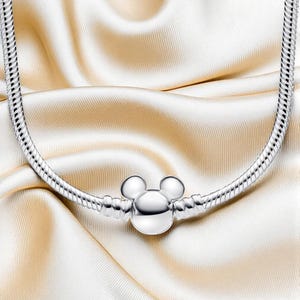 K&ouml;nnte beinhalten: Eine silberne Halskette mit einem Mickey-Mouse-Anh&auml;nger. Die Halskette hat ein Schlangenketten-Design und der Anh&auml;nger ist eine dreidimensionale Darstellung von Mickey Mouses Kopf. Pr&auml;sentiert auf cremefarbenem Satin.