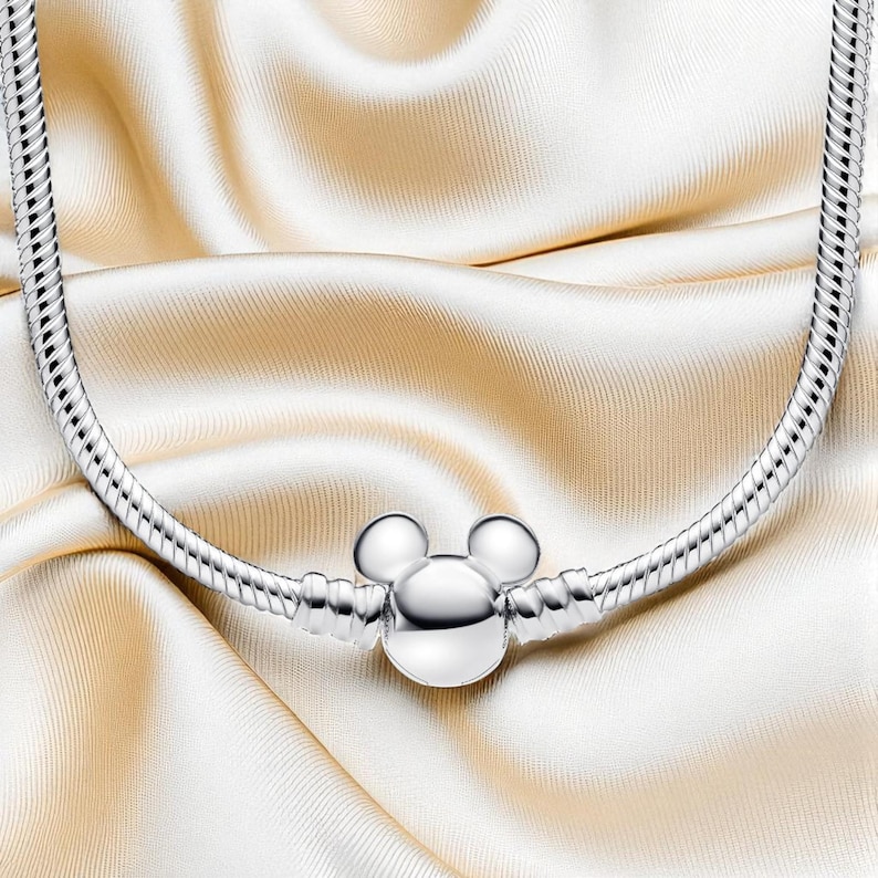 K&ouml;nnte beinhalten: Eine silberne Halskette mit einem Mickey-Mouse-Anh&auml;nger. Die Halskette hat ein Schlangenketten-Design und der Anh&auml;nger ist eine dreidimensionale Darstellung von Mickey Mouses Kopf. Pr&auml;sentiert auf cremefarbenem Satin.