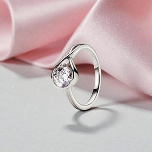 May include: A silver ring with a teardrop-shaped setting holding a clear, sparkling gemstone. The ring band has a twisted design, and the gemstone is the focal point. The background features a soft pink fabric.
