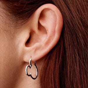 Pu&ograve; includere: Primo piano di un orecchino color argento a forma di mela stilizzata. L'orecchino &egrave; a cerchio, con il design della mela che pende sotto il lobo dell'orecchio. Lo sfondo mostra un orecchio e capelli castani.