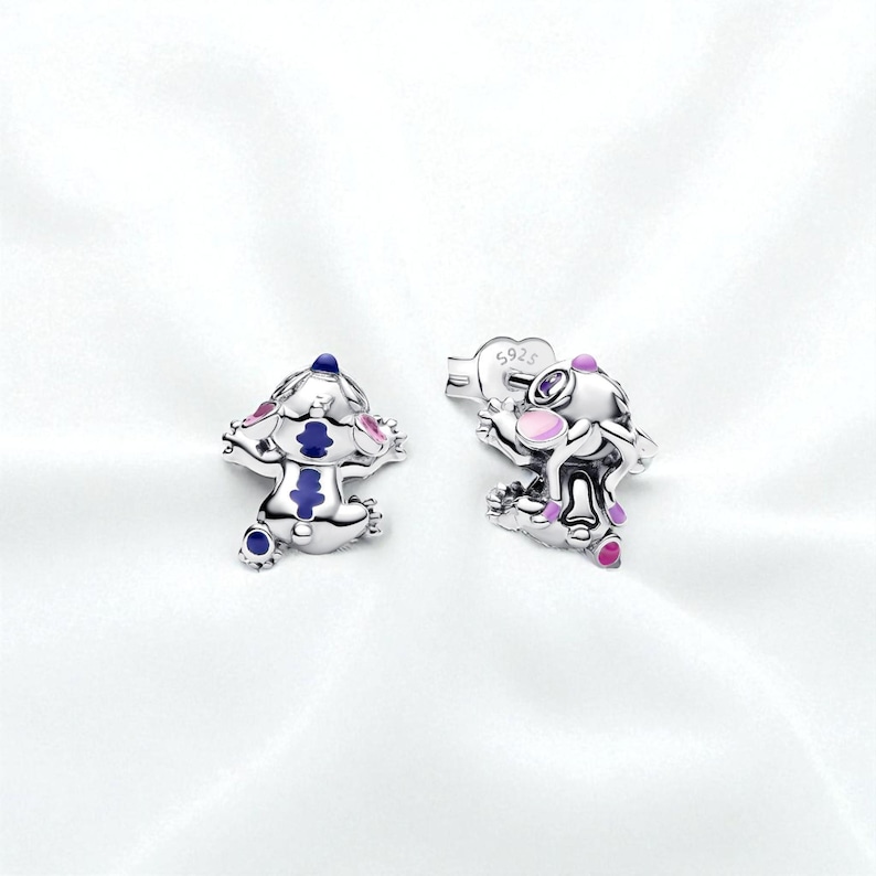 Pu&ograve; includere: Un paio di orecchini a lobo argentati con un design a personaggio dei cartoni animati. Gli orecchini hanno dettagli in smalto blu, rosa e viola. Sono esposti su uno sfondo bianco.