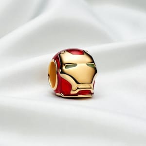May include: A charm depicting the Iron Man helmet in red and gold. The charm has a detailed design of the helmet's face. It is likely made of metal and is designed to be added to a bracelet or necklace, a popular accessory.