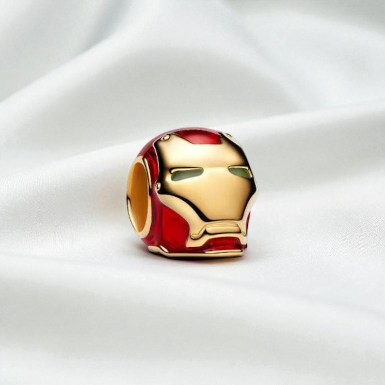 May include: A charm depicting the Iron Man helmet in red and gold. The charm has a detailed design of the helmet's face. It is likely made of metal and is designed to be added to a bracelet or necklace, a popular accessory.