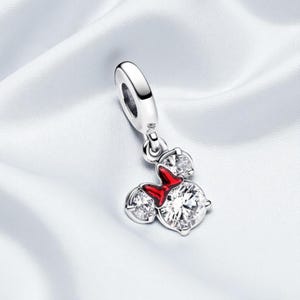 May include: A silver charm with a Minnie Mouse design. The charm features three clear, sparkling cubic zirconia stones and a red enamel bow. The charm is designed to be added to a bracelet or necklace.