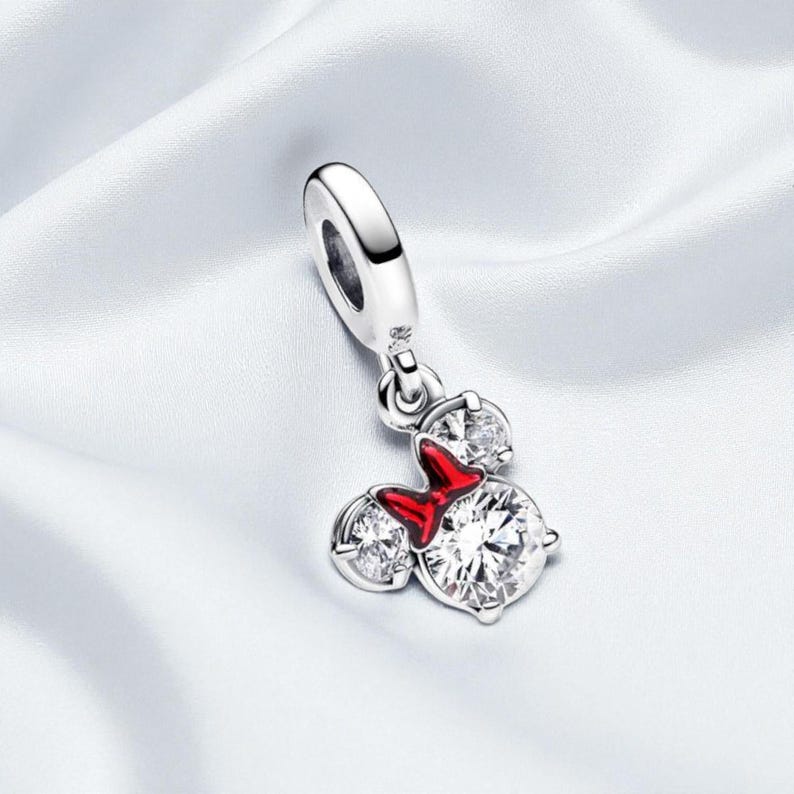 May include: A silver charm with a Minnie Mouse design. The charm features three clear, sparkling cubic zirconia stones and a red enamel bow. The charm is designed to be added to a bracelet or necklace.