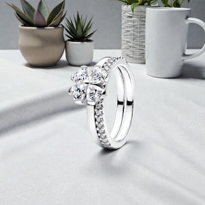 Puede incluir: Un anillo plateado con un diseño de trébol de cuatro hojas, con piedras preciosas claras y brillantes. La banda está adornada con piedras más pequeñas y claras. El anillo se muestra sobre una superficie blanca, con plantas y una taza blanca en el fondo.