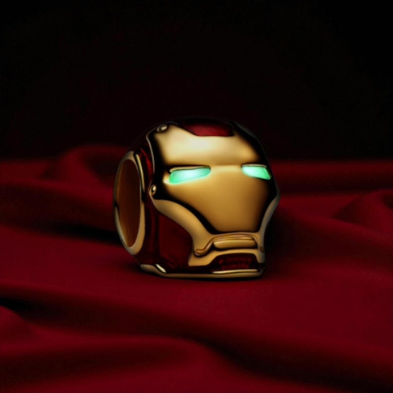 May include: A gold and red Iron Man helmet ring with glowing green eyes. The ring is set against a dark red, textured background. The ring is a detailed replica of the superhero's iconic helmet. The ring is a detailed replica.