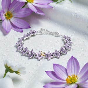 Op de afbeelding: Een zilveren armband versierd met lavendelkleurige bloemvormige edelstenen. De armband heeft een delicate ketting en sluiting, met een klein gouden accent. De sieraden worden gepresenteerd op een wit, getextureerd oppervlak, omringd door paarse bloemen.