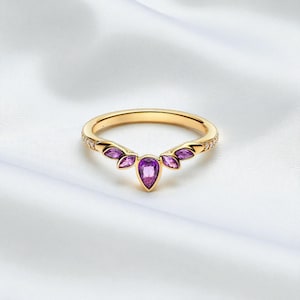 Puede incluir: Un anillo dorado con una piedra central púrpura en forma de pera y piedras púrpuras más pequeñas en un diseño en forma de abanico. La banda del anillo está adornada con pequeñas piedras preciosas transparentes. El anillo está sobre un fondo blanco.