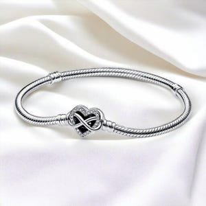 May include: A silver bracelet with a heart-shaped charm featuring an infinity symbol. The charm is encrusted with small, clear crystals. The bracelet has a snake chain design and is displayed on a white, silky background.
