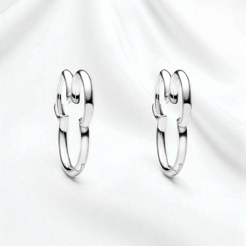 Pu&ograve; includere: Un paio di orecchini a cerchio color argento. Ogni orecchino presenta un design unico con due anelli ad incastro, creando un look moderno ed elegante. Gli orecchini sono esposti su uno sfondo bianco.