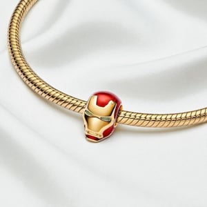 May include: A gold charm bracelet featuring a red and gold Iron Man helmet charm. The charm has green eyes and a gold faceplate. The bracelet is set against a white backdrop.