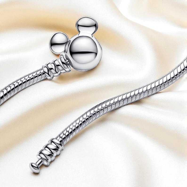 K&ouml;nnte beinhalten: Ein silbernes Armband mit einem Mickey Mouse-Anh&auml;nger. Der Anh&auml;nger hat einen runden Kopf und zwei kleine runde Ohren. Das Armband hat ein Schlangenketten-Design und wird auf einer wei&szlig;en Satin-Oberfl&auml;che gezeigt.
