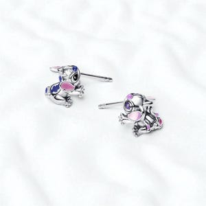 Pu&ograve; includere: Un paio di orecchini a bottone argentati con un design a personaggio dei cartoni animati. Gli orecchini hanno dettagli in smalto blu, rosa e viola. Sono presentati su uno sfondo bianco.