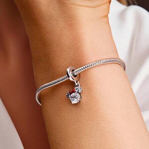 May include: A silver charm bracelet with a Minnie Mouse charm. The charm features a clear, round gemstone with a red bow and two smaller clear gemstones. The bracelet is a snake chain style.