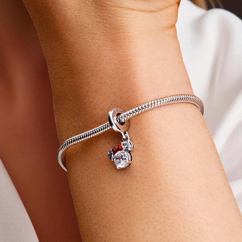 May include: A silver charm bracelet with a Minnie Mouse charm. The charm features a clear, round gemstone with a red bow and two smaller clear gemstones. The bracelet is a snake chain style.